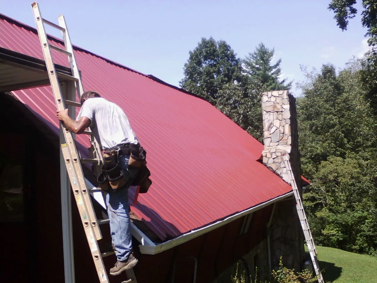 Roofer on a ladder beside a red metal roof and stone chimney — Soffit & Fascia Repair by Roofing Brothers in Blountstown